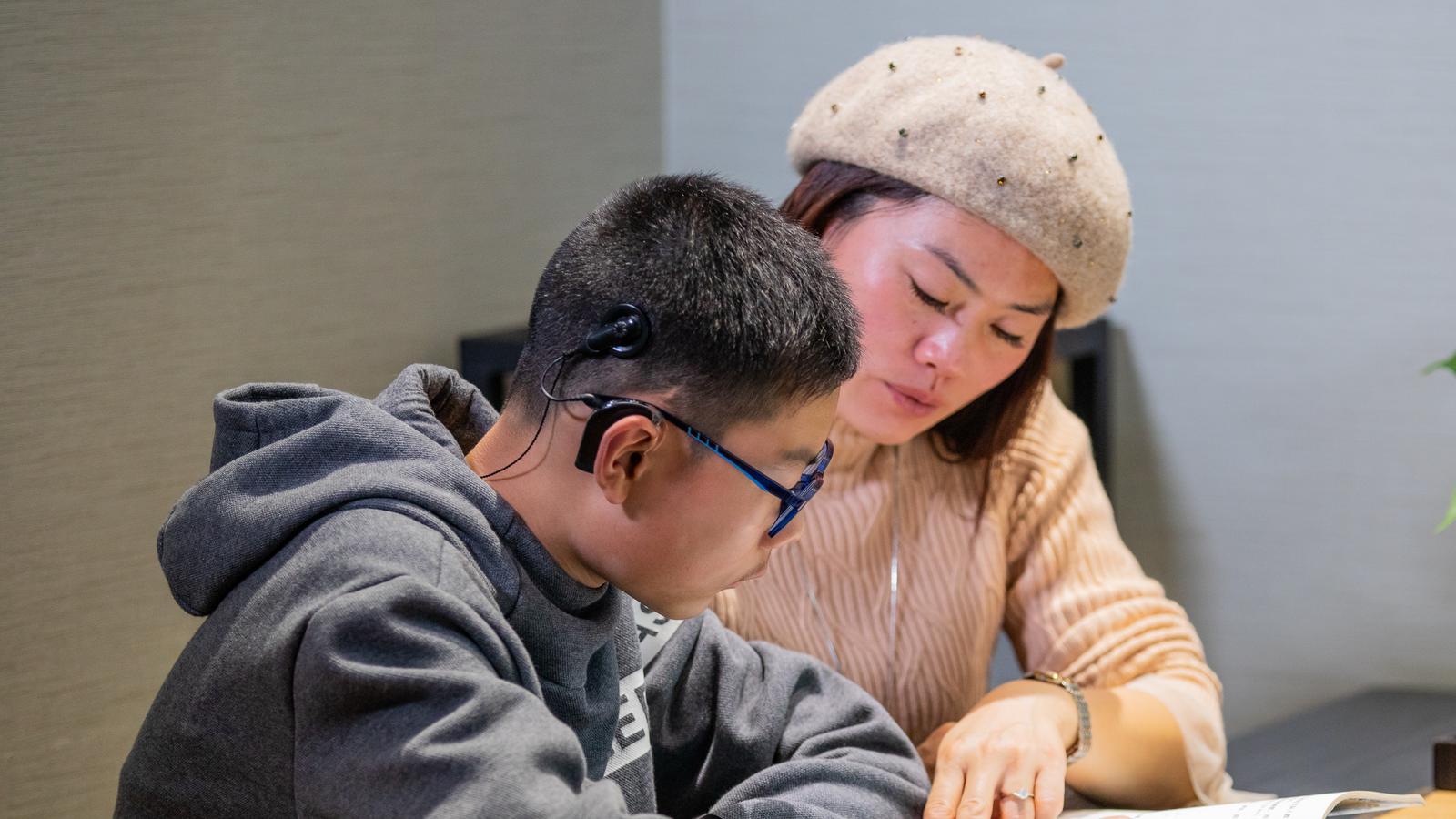 A woman sits with a boy wearing a cochlear implant