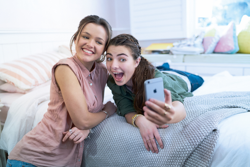 Two friends pose for a photo with an iPhone in a bedroom