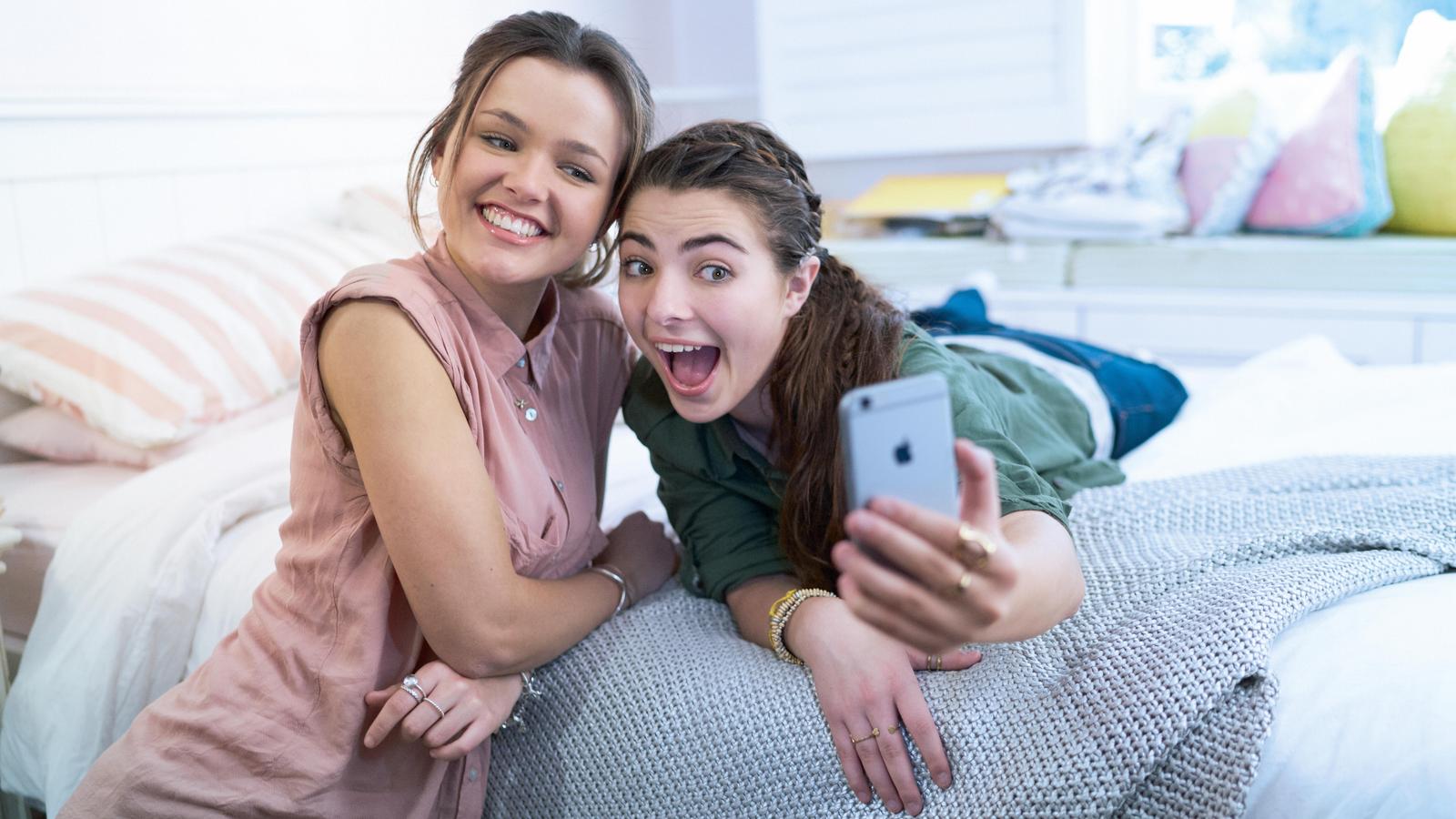 Dvě kamarádky v ložnici pózují s iPhonem pro fotografa
