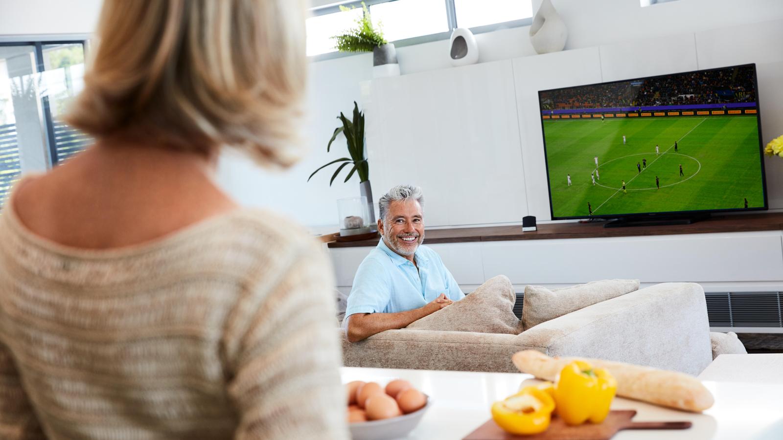 Bărbat adult urmărind un meci de fotbal la TV cu Cochlear Wireless TV Streamer (streamer TV)