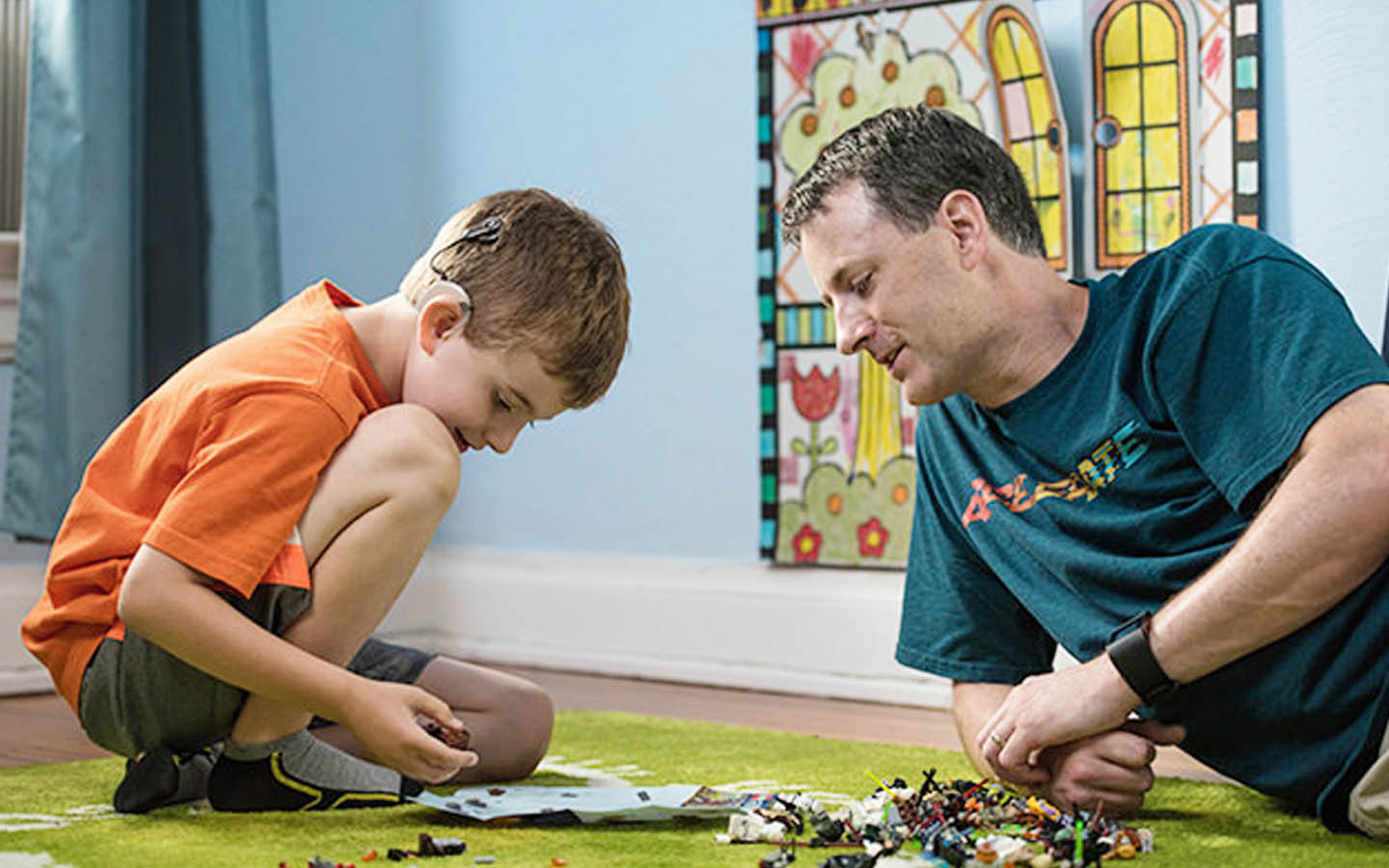 Egy padlón játszó, cochleáris implantátumot viselő kisfiú, akit édesapja néz