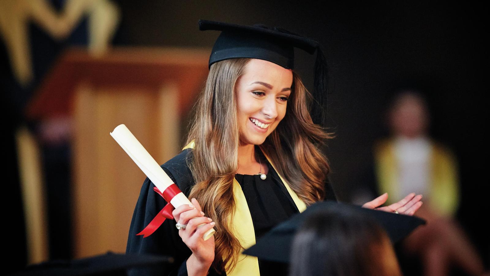A woman wearing a mortarboard and gown holds a diploma