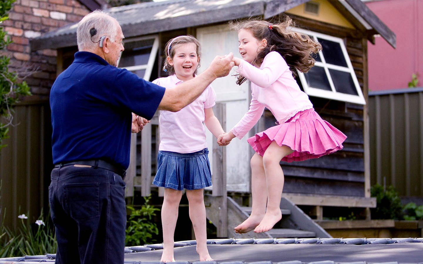 Un paciente con implante coclear sostiene las manos de sus dos nietas mientras saltan en una cama elástica