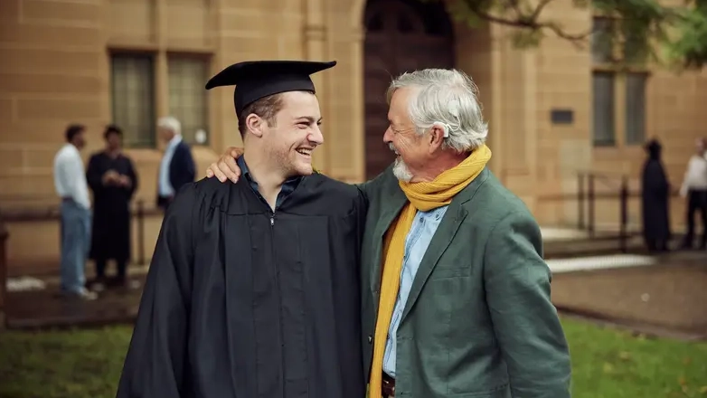 Older man wearing a Cochlear Nucleus Nexa Implant smiles with a young graduate after a graduation ceremony.