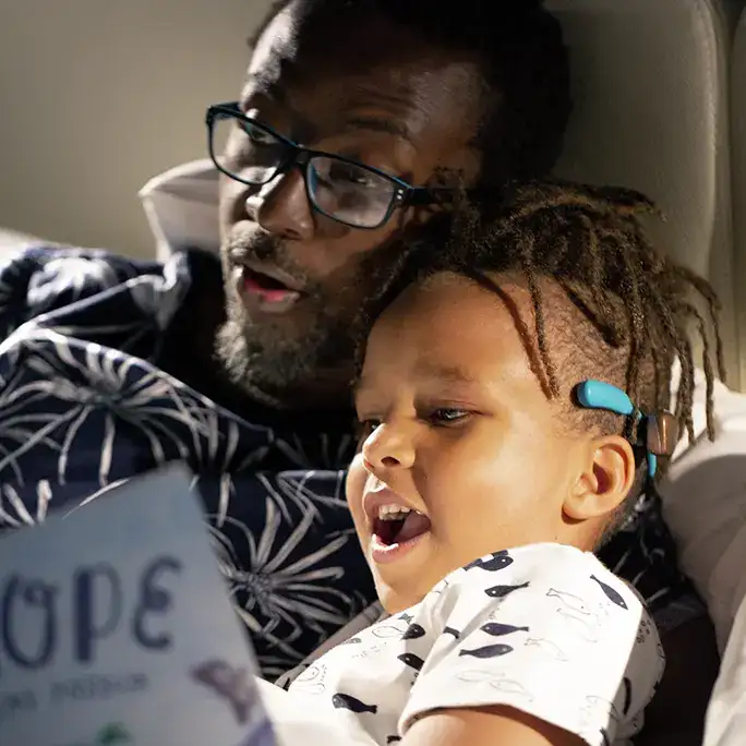 Ein kleiner Junge mit einem türkisen SoundArc schaut gemeinsam mit seinem Vater im Bett ein Buch an.