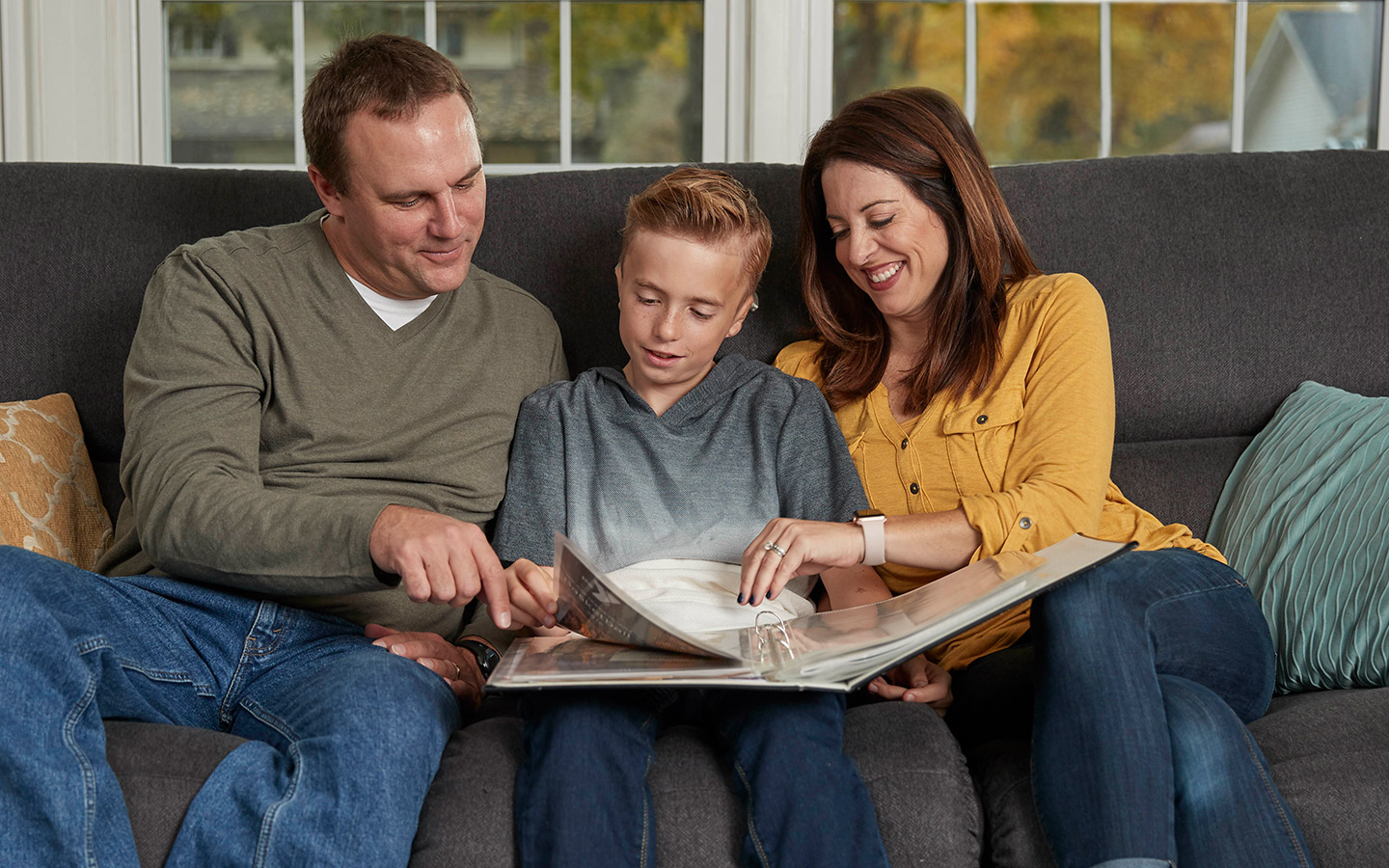A mother and father sit on a couch with son, wearing a Cochlear implant, and listen to him read