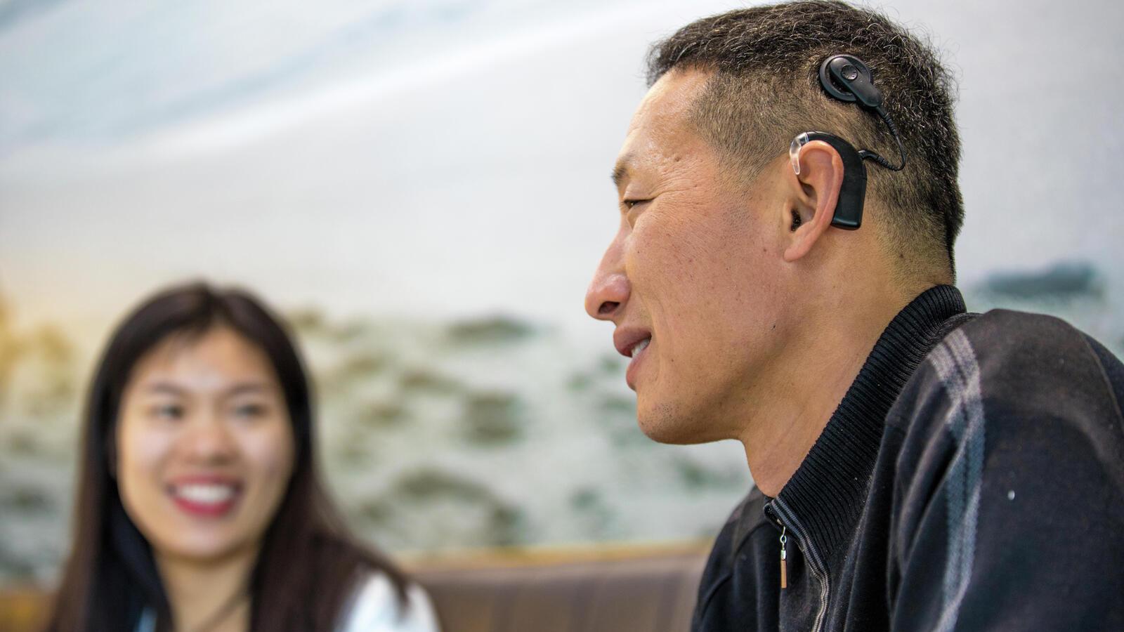 A mother and father sit on a couch with son, wearing a Cochlear implant, and listen to him read