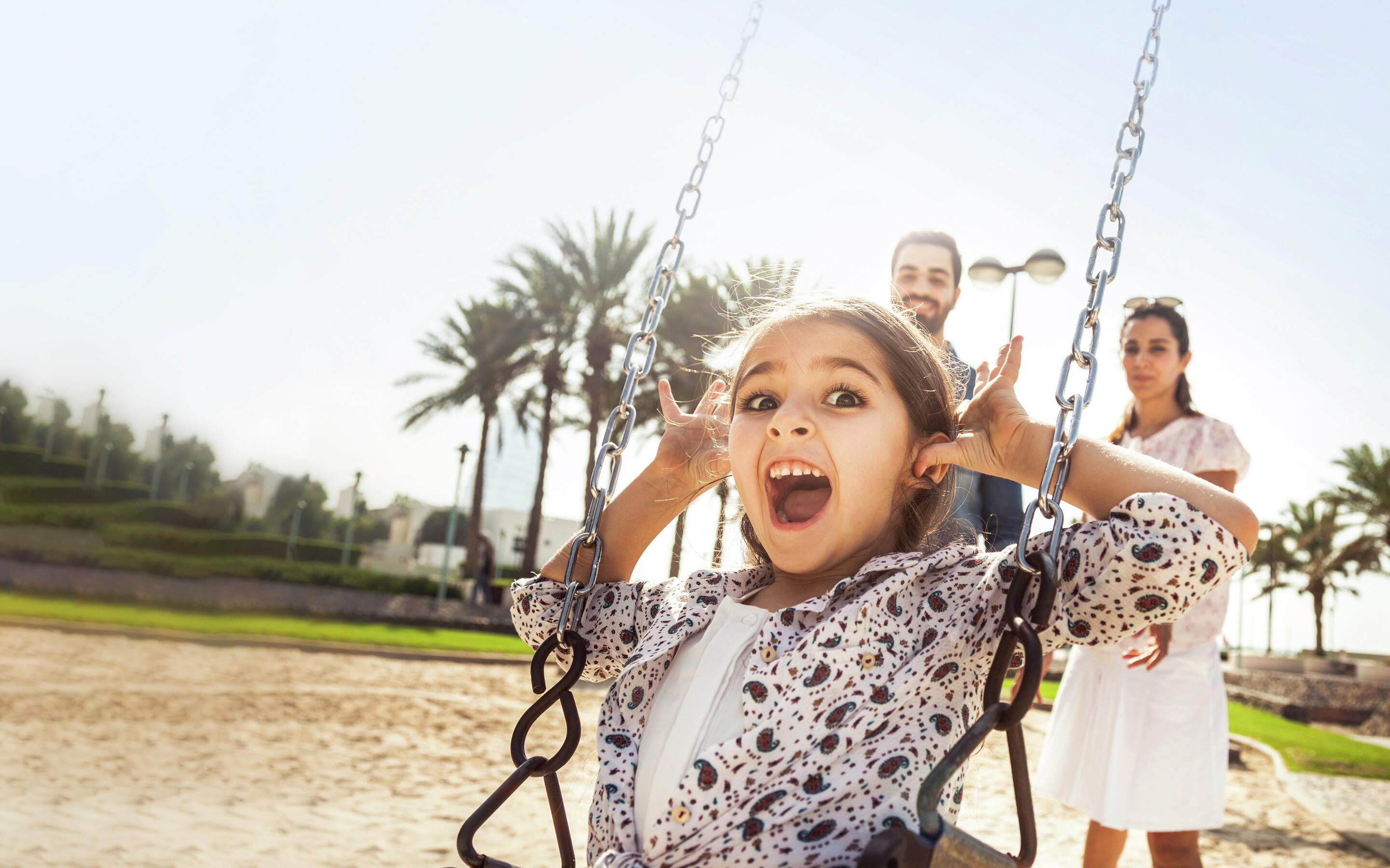 A young girl swings at playground with her parents in the background